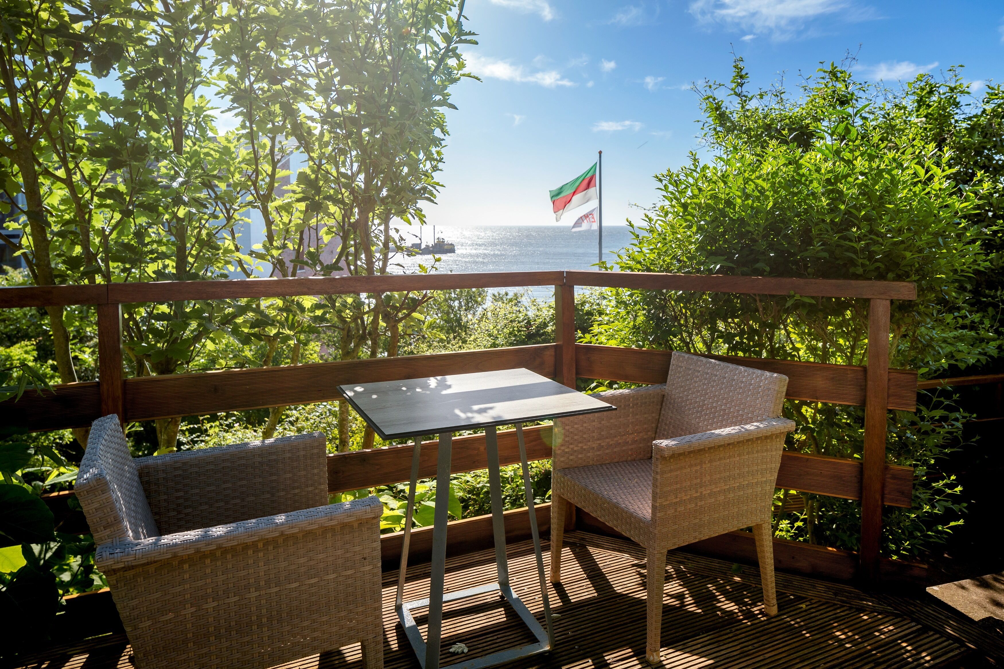 Una terraza de madera con mobiliario de mimbre y una mesa, con vista a un cuerpo de agua e una bandera.