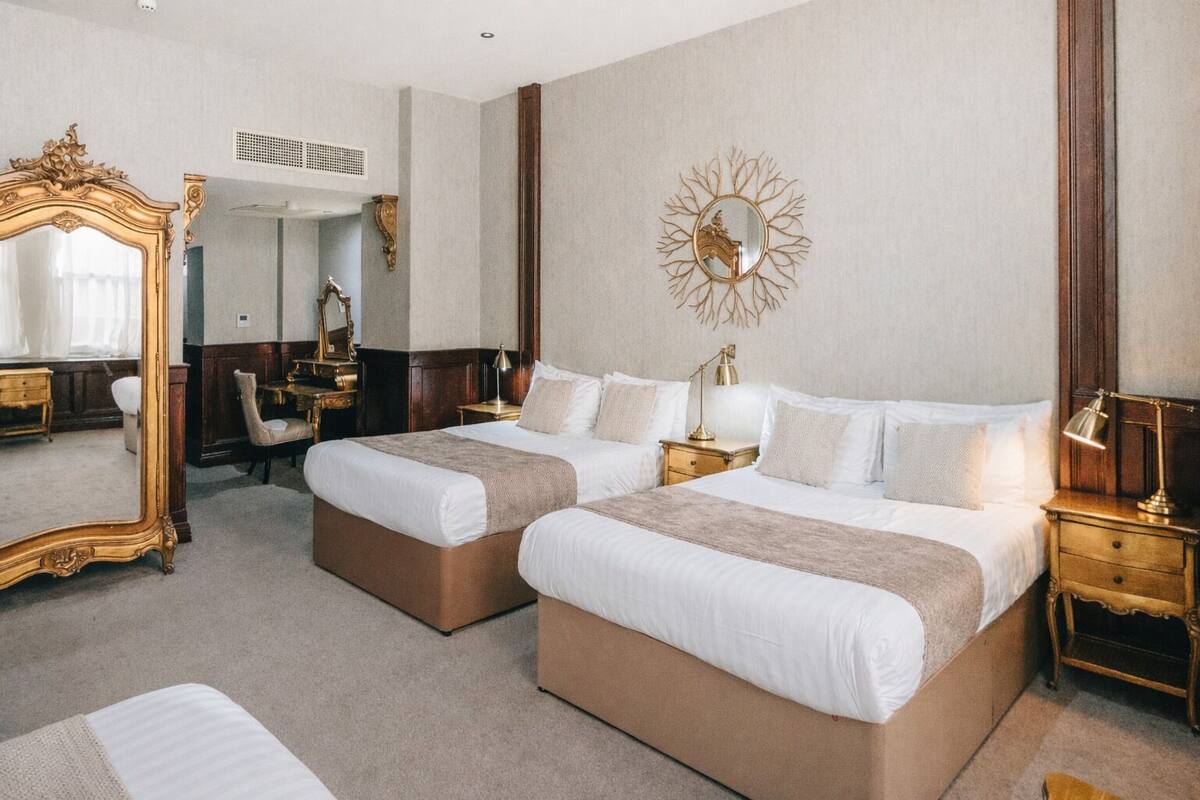 A hotel room with two beds, a large ornate mirror, a wall-mounted decorative piece, and a desk with a chair.