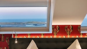 Una habitación con vista al mar a través de un tragaluz, una pared roja con estampado de langosta y un cabecero tapizado en negro.