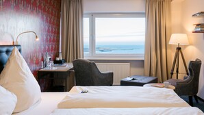 Habitación de hotel con una cama grande, un escritorio, dos sillas, una lámpara y vista al mar a través de una ventana.