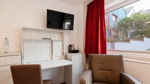 A hotel room with a white desk, a brown chair, a television mounted on the wall, and a window with red curtains. - Hotel Felsen-Eck (Helgoland)