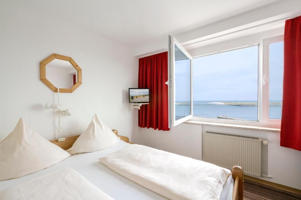 A hotel room with a bed, a window with red curtains, a wall-mounted mirror, and a view of the sea.