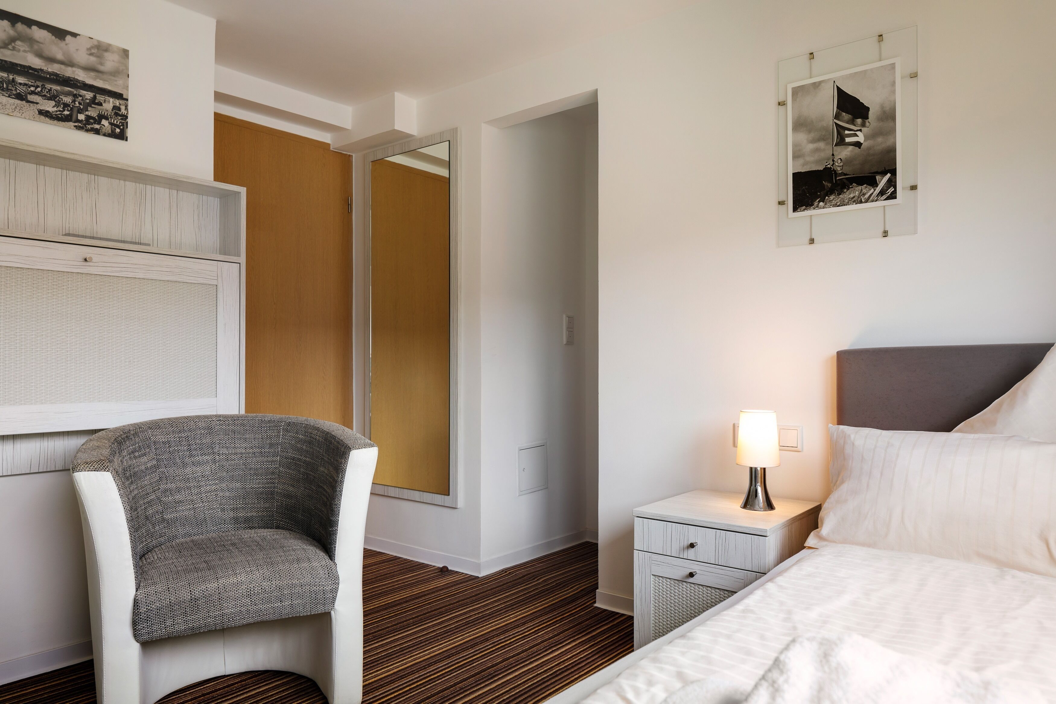 Een hotelkamer met een wit bed, een grijze fauteuil, een nachtkastje met een lamp en een ingelijst schilderij aan de muur.