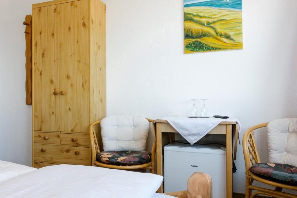 Una habitación con un armario de madera, dos sillones tapizados con cojines, una mesita con una copa y una botella, y una cama con ropa de cama blanca.