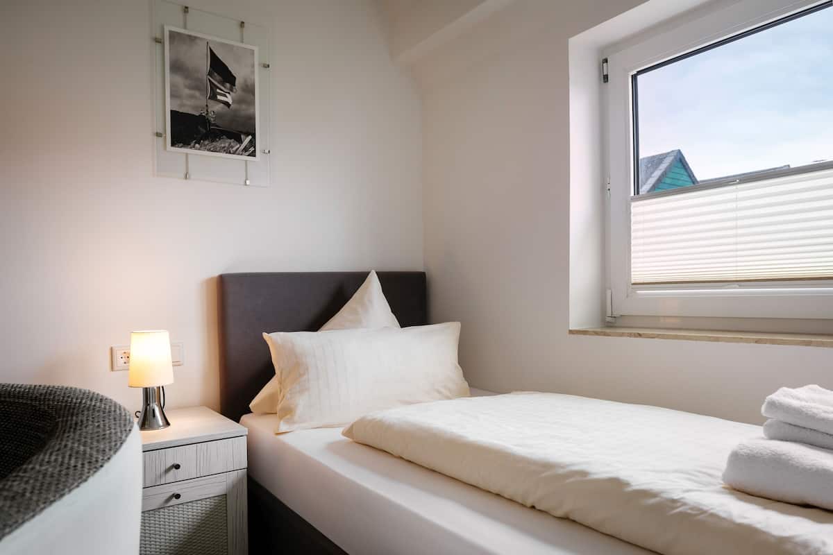Cama bem arrumada com lençóis brancos e travesseiro, luminária de cabeceira, mesa de cabeceira pequena com uma luminária e um quadro emoldurado na parede.