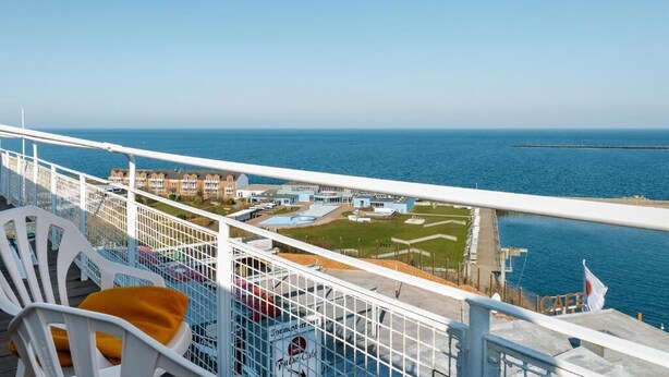 A balcony with white chairs and a yellow cushion, overlooking a coastal area with buildings and a pier. - Hotel Felsen-Eck (Helgoland)