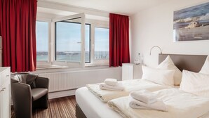 A hotel room with a bed, a chair, a desk, a window with a view of the sea, and a painting on the wall. - Hotel Felsen-Eck (Helgoland)