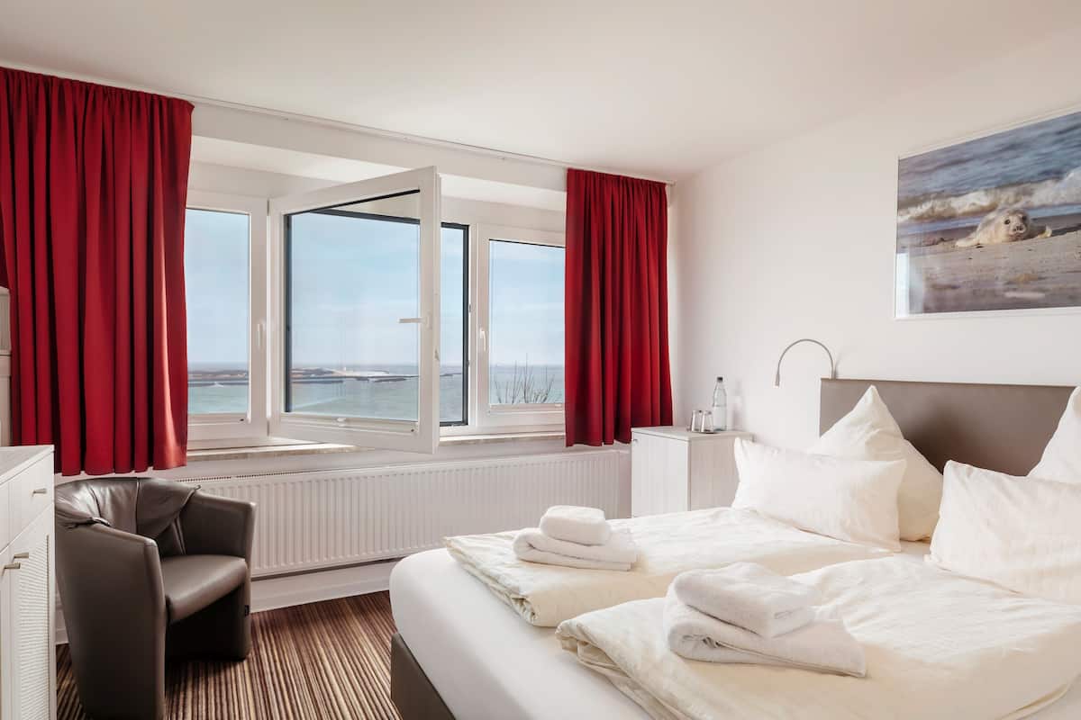 Quarto de hotel com cama, uma cadeira, uma escrivaninha, janela com vista para o mar e um quadro na parede.