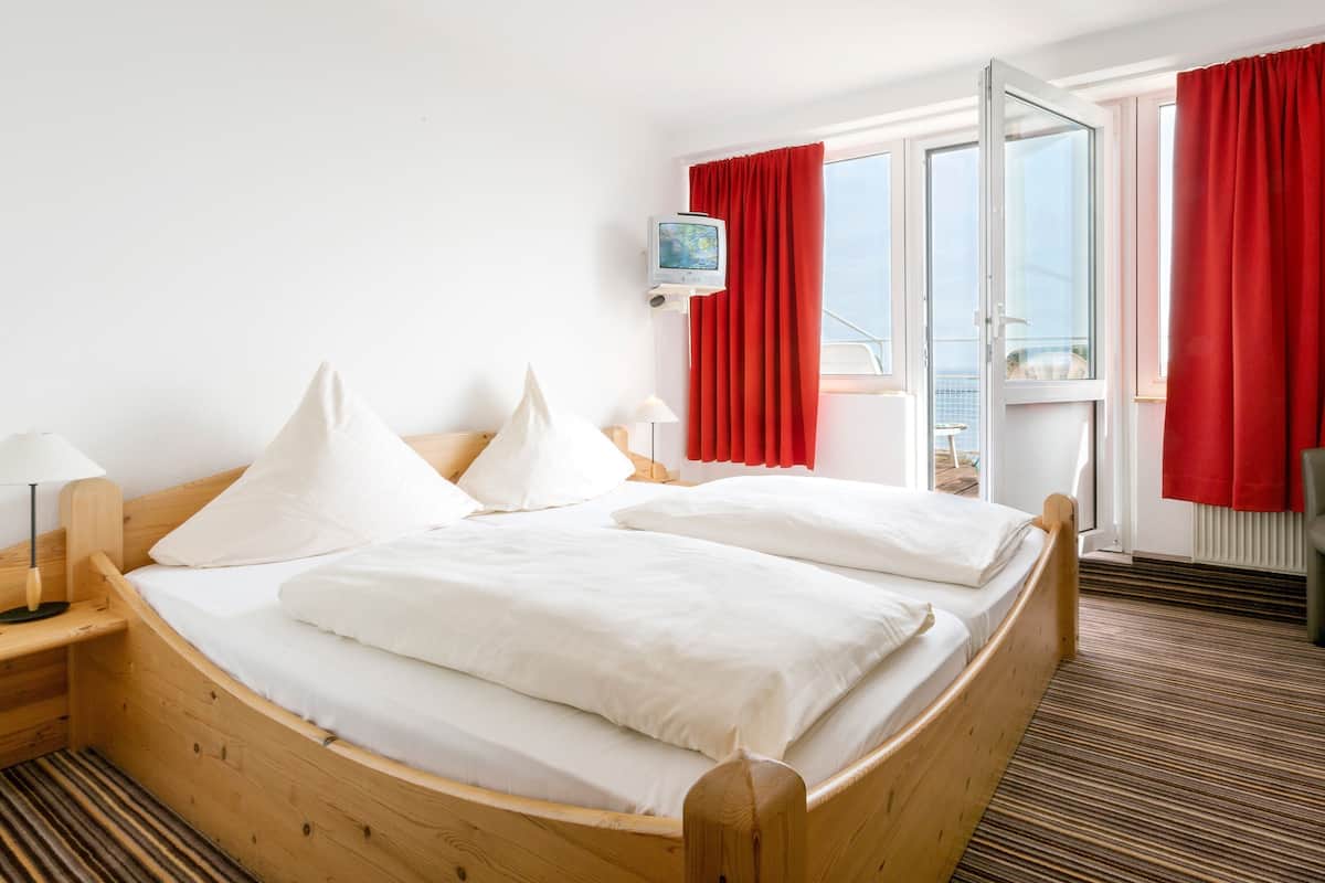 Uma cama de madeira com roupa de cama branca num quarto com vista para o mar através de uma porta de vidro.
