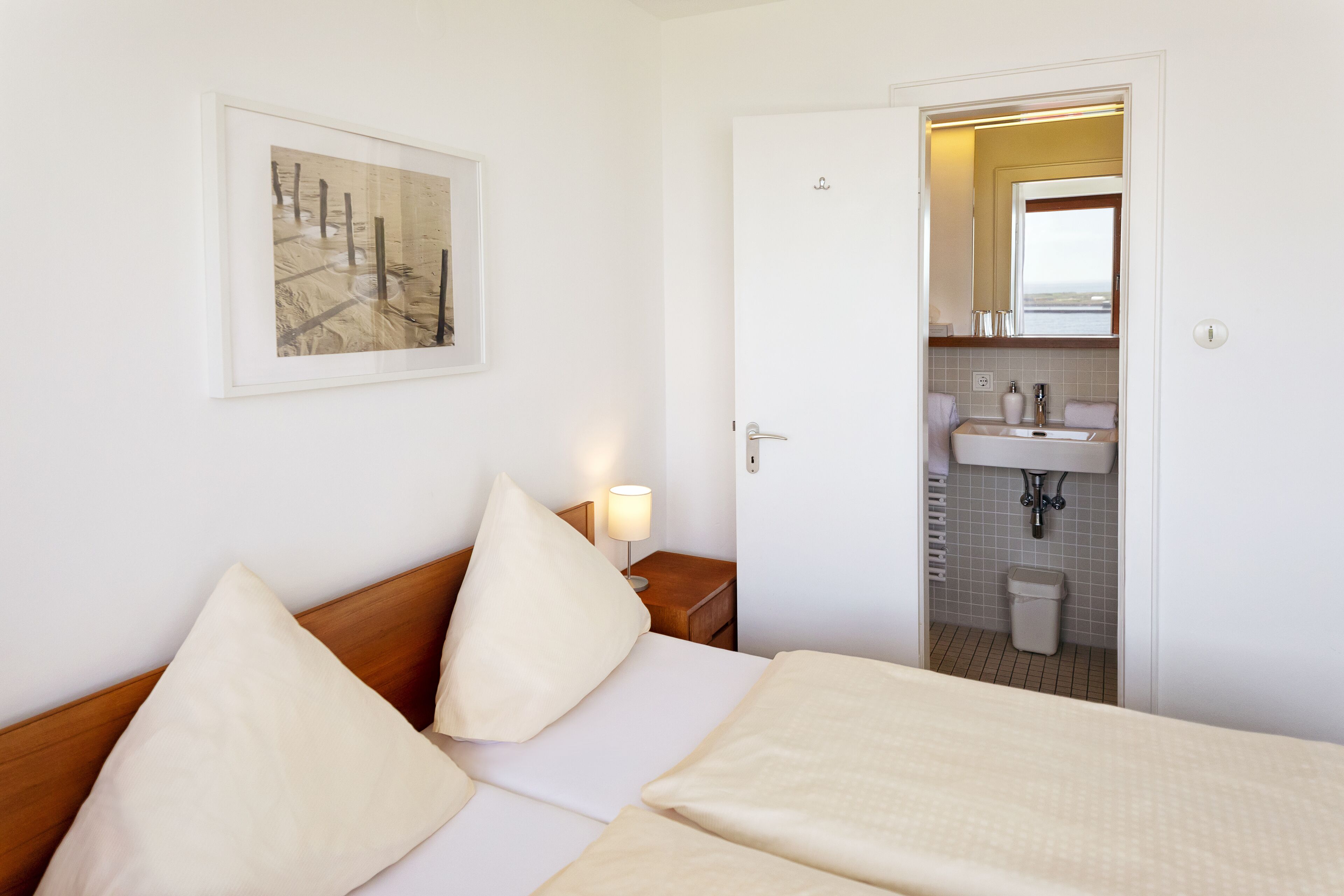 A hotel room with a bed, pillows, a bedside table with a lamp, a framed picture on the wall, and a view into the bathroom.