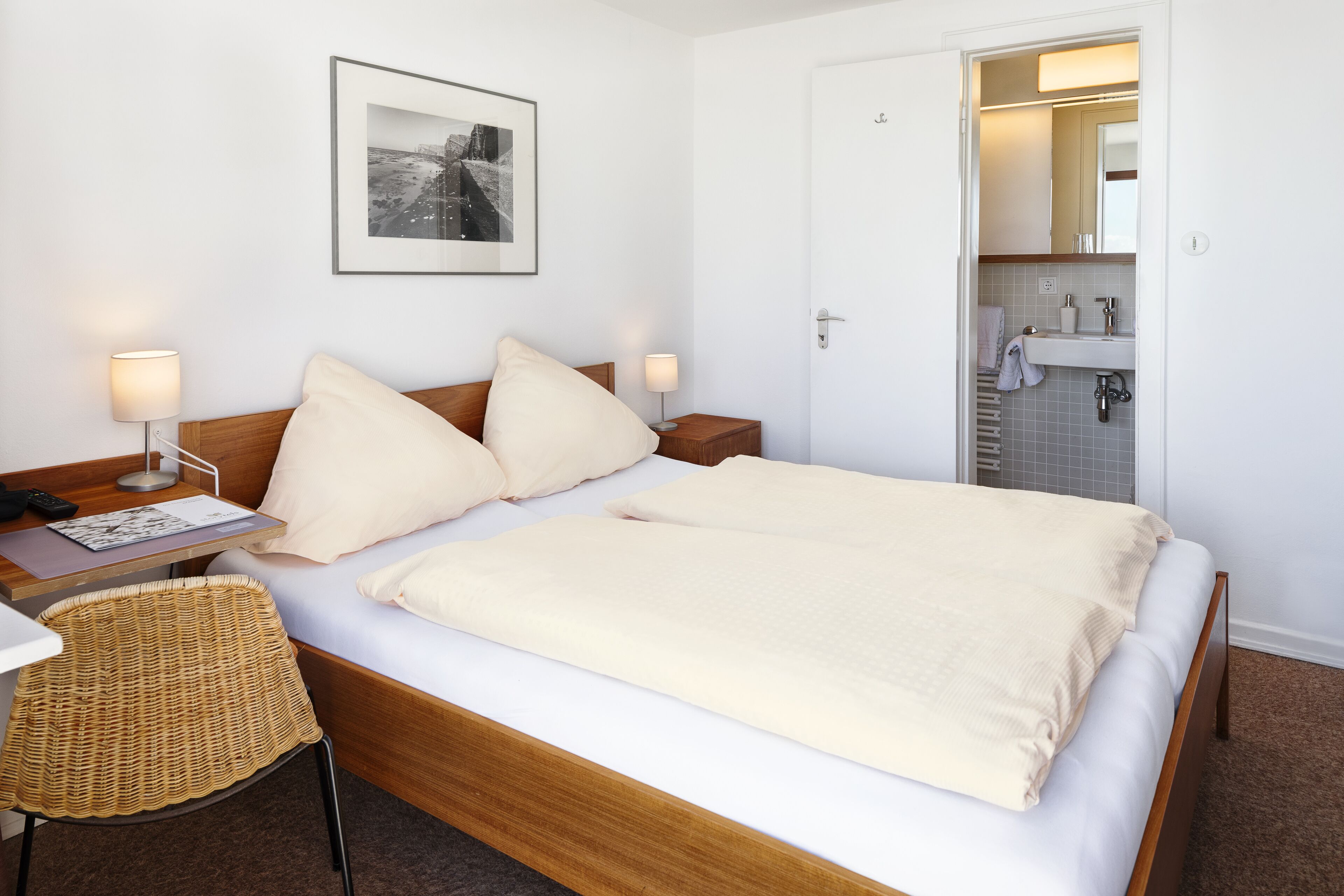 Habitación de hotel con cama, mesitas de noche, una silla, un cuadro en la pared y un baño visible a través de una puerta abierta.