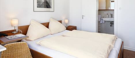 Habitación de hotel con cama, mesitas de noche, una silla, un cuadro en la pared y un baño visible a través de una puerta abierta.