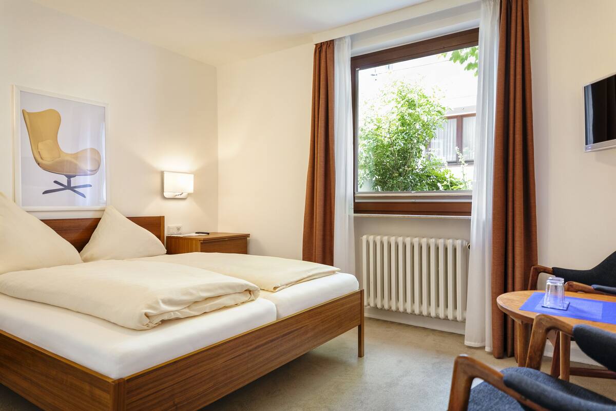 Quarto de hotel com cama, mesa de cabeceira, luminária, janela com cortinas e vista para o verde lá fora.