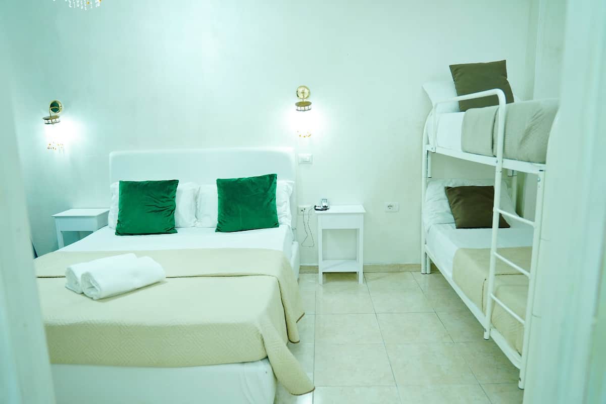Quarto de hotel com uma cama de solteiro, uma cama beliche, uma mesinha de cabeceira e um ponto de luz fixado na parede.