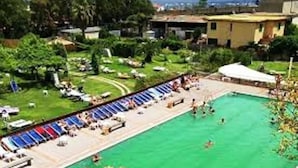 Una piscina amb gandules i zona coberta, envoltada de vegetació i edificis.