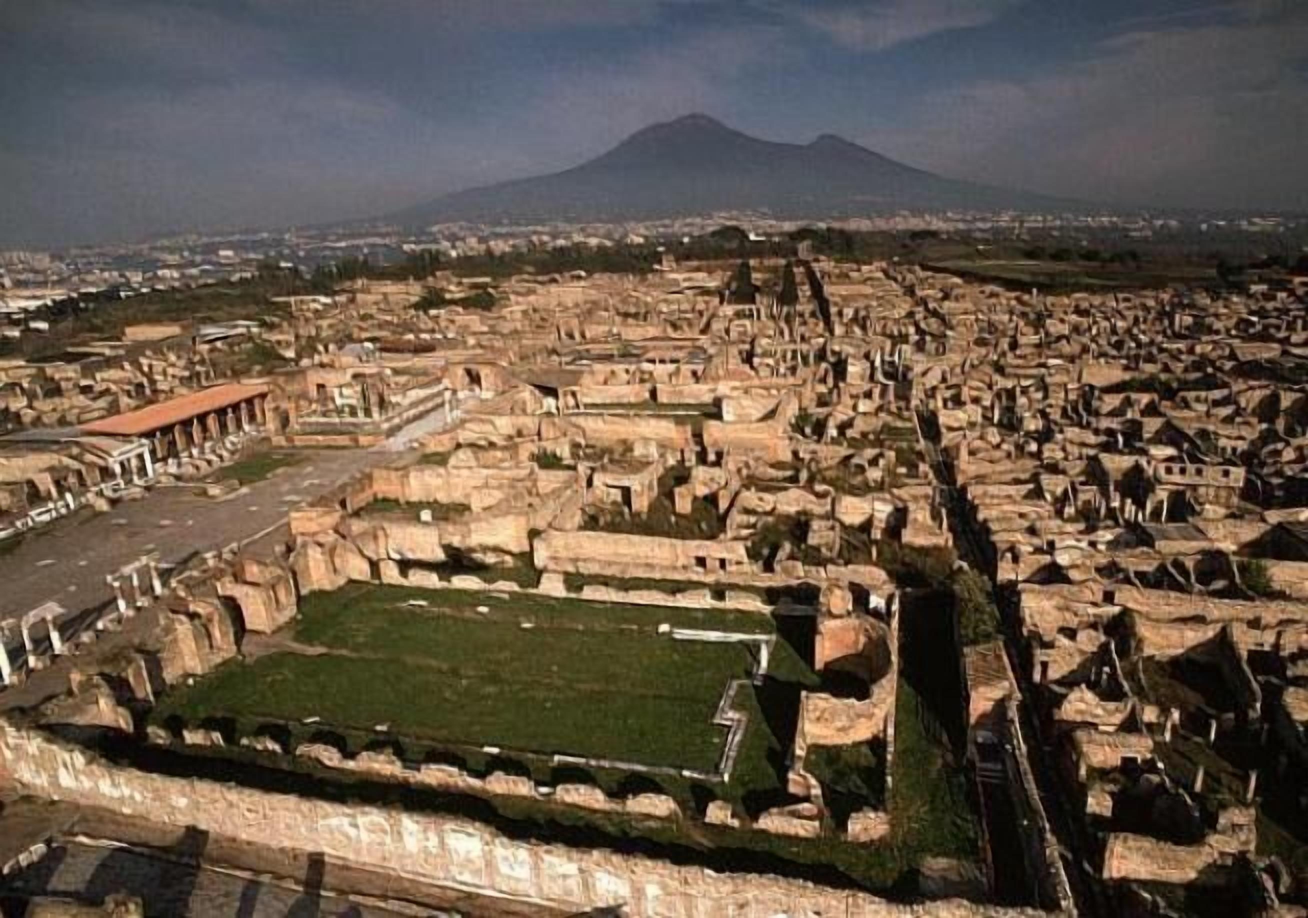 An aerial view of an ancient city with ruins and a large open green space in the foreground.