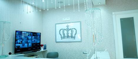 Uma área de recepção com televisão exibindo imagens de segurança, um quadro com uma coroa e um candelabro.
