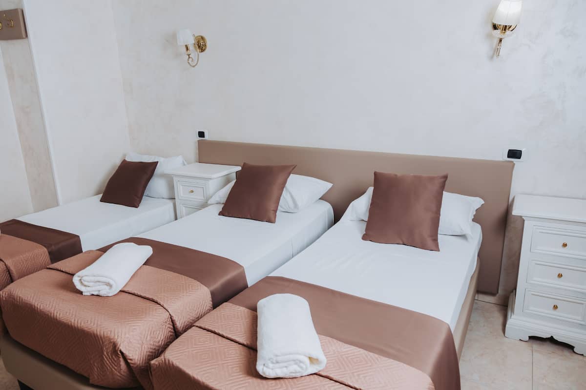 Quarto de hotel com três camas, cada uma com cabeceira marrom, roupa de cama branca e toalha enrolada no rodapé. Há duas mesas de cabeceira com luminárias, e um criado-mudo branco com uma luminária.