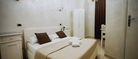 A neatly made bed with a beige bedspread, two brown pillows, and a folded white towel. There is a white wardrobe with a mirror and a small bedside table with a lamp.