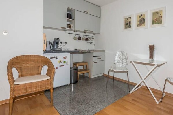 A compact kitchen with white cabinets, a wicker chair, a small dining table, and a trash bin.