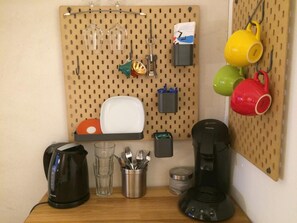 Un coin cuisine avec un mur équipé d’un panneau perforé, une machine à café, une bouilloire, un verre, un porte-ustensiles et des tasses colorées.
