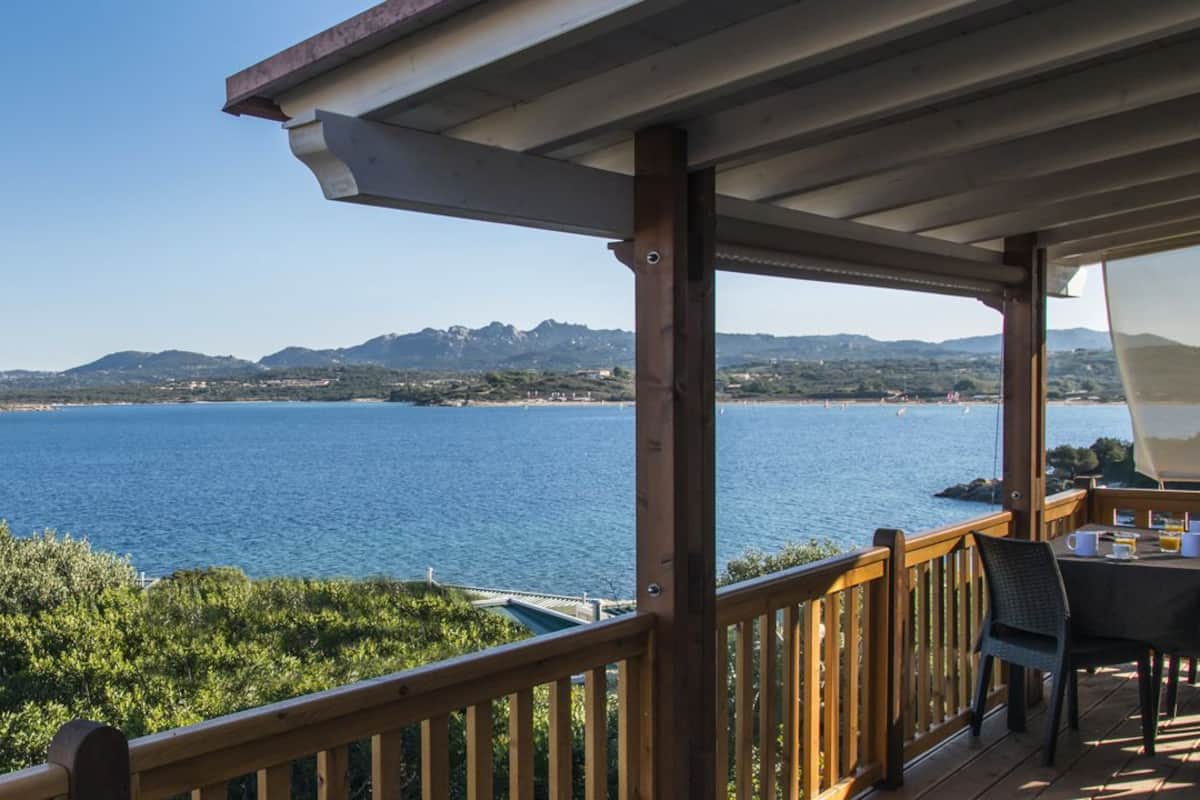 A wooden deck with a table and chairs overlooking a body of water and distant mountains.