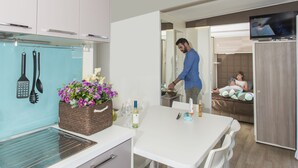 Cozinha moderna com área de jantar, televisão na sala e quarto com cama e uma pessoa sentada.