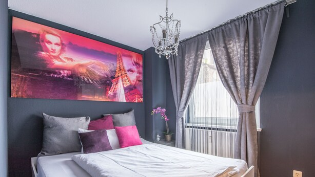 Een slaapkamer met een bed, een kroonluchter, een raam met gordijnen en een schilderij van de Eiffeltoren.