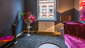 Een kamer met een radiator, een raam met uitzicht op een gebouw, een vaas met bloemen en een rode bank.