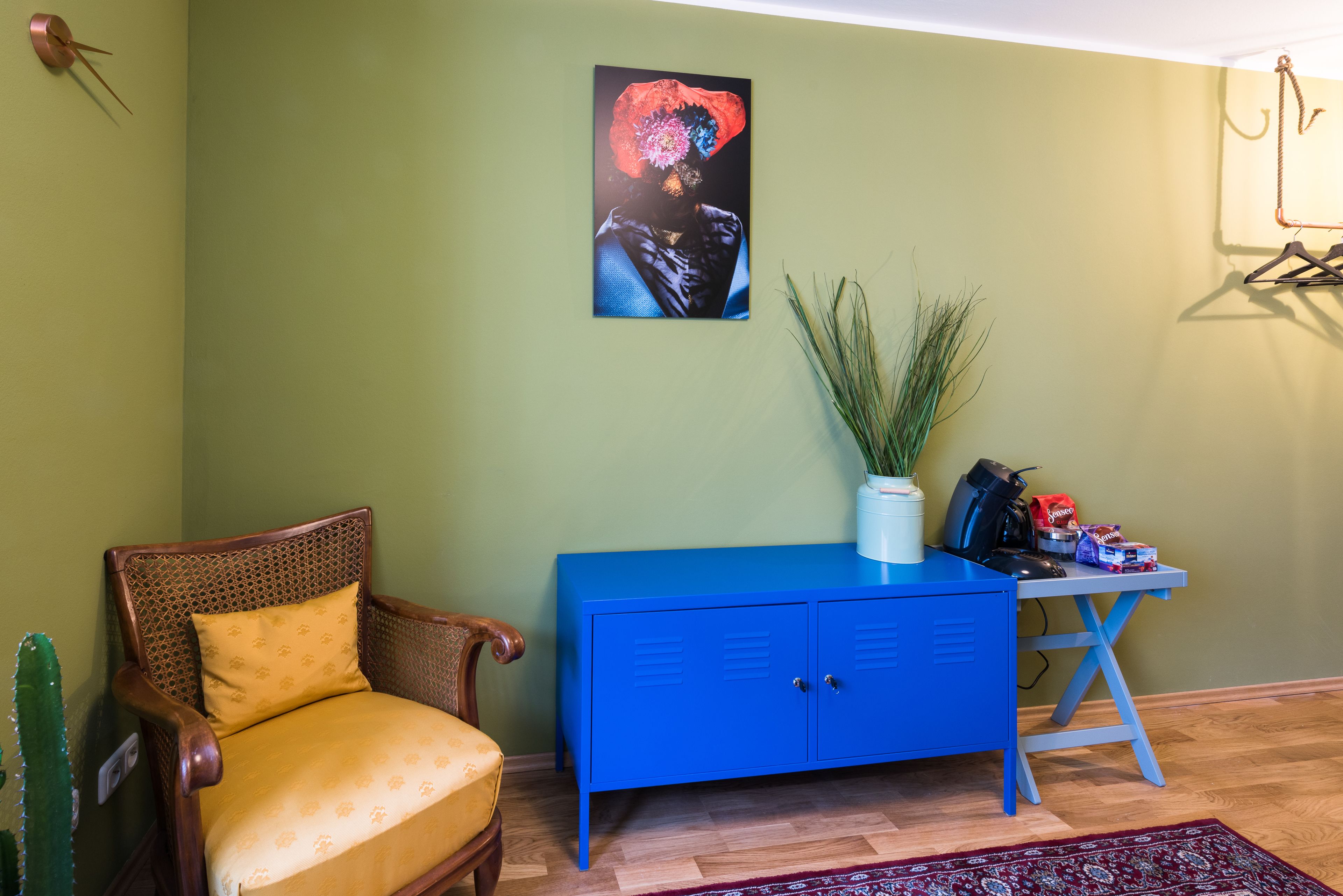 Een kamer met een houten stoel, een blauwe kast, een cactus en een schilderij aan de muur.