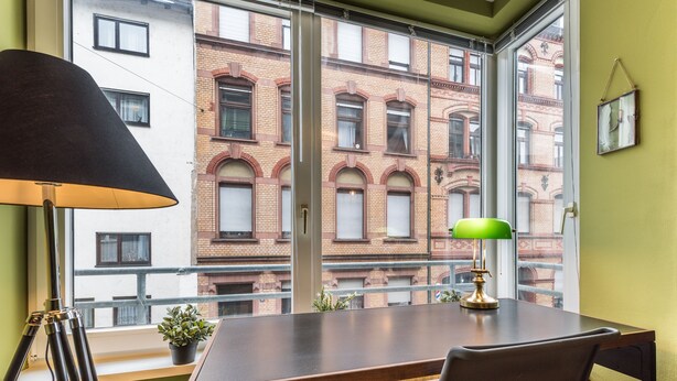Une pièce avec une grande fenêtre, un bureau, une chaise, une lampe et une plante en pot.