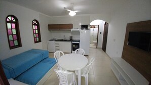 Un espacio habitable compacto con kitchenette, una zona de comedor con mesa y sillas, un sofá azul y un televisor montado en la pared.