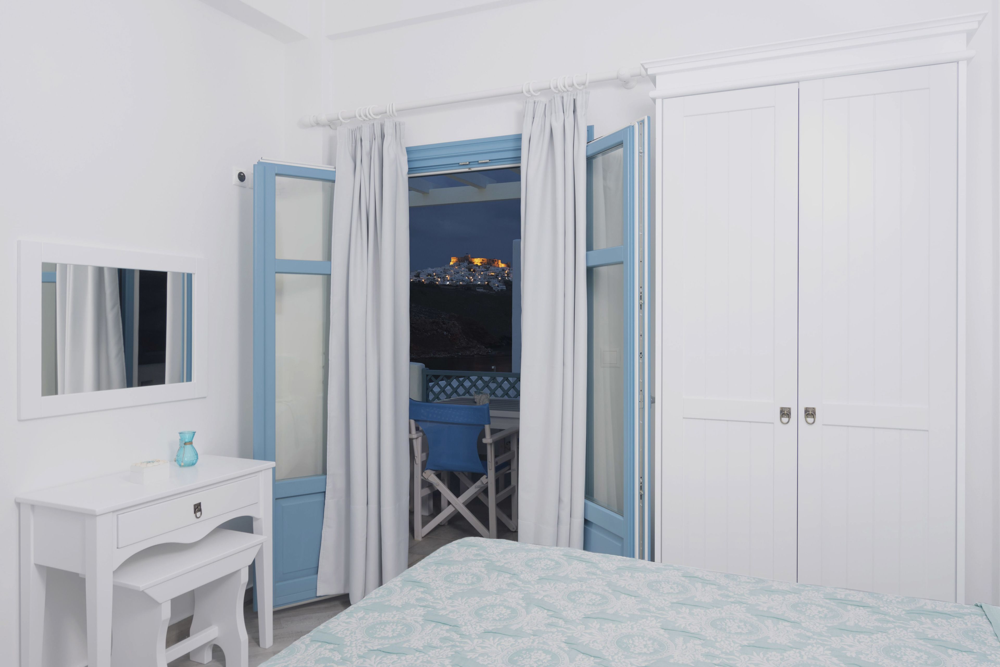 A bedroom with a bed, a desk, and a chair. There is a wardrobe with a mirror and a window with curtains.