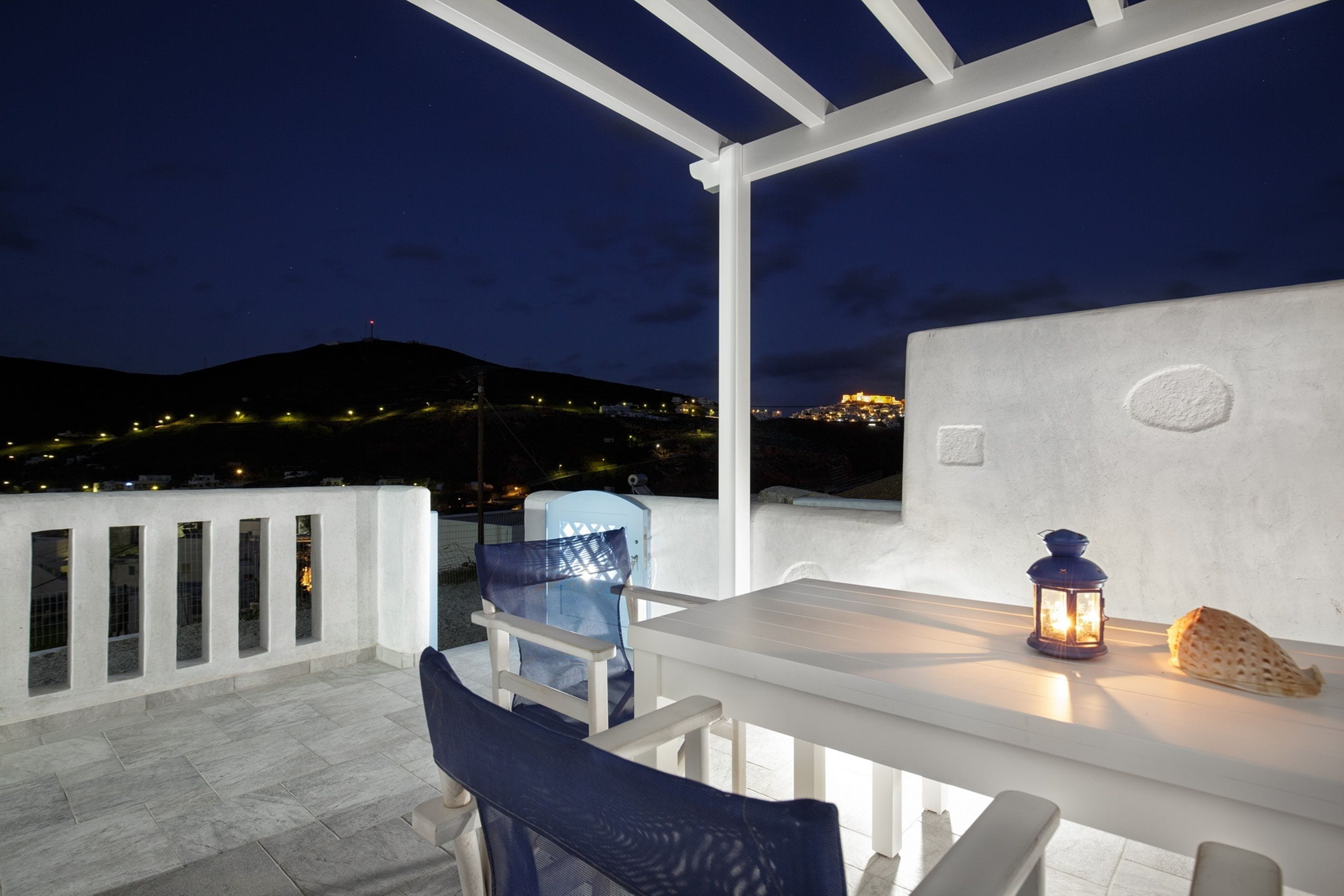 Un patio al aire libre con una mesa y sillas blancas, un farol y vistas de un paisaje urbano nocturno.