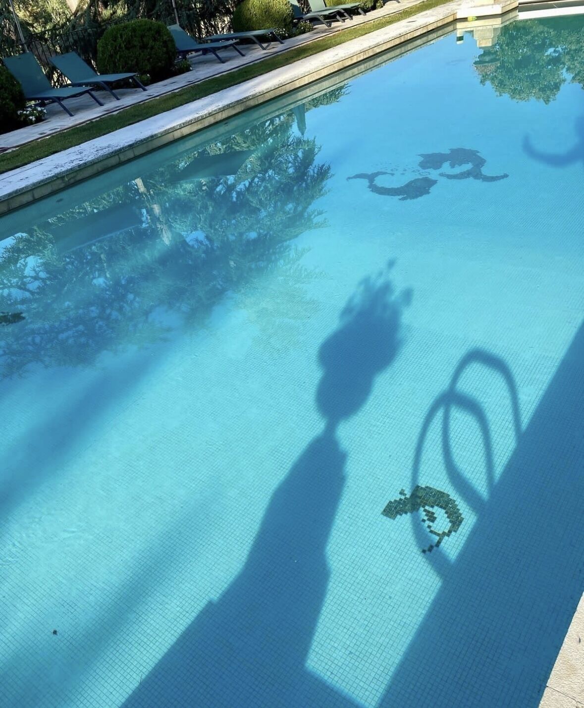 Une piscine aux eaux cristallines, bordée de carreaux, et on aperçoit le reflet d’une personne dans l’eau.