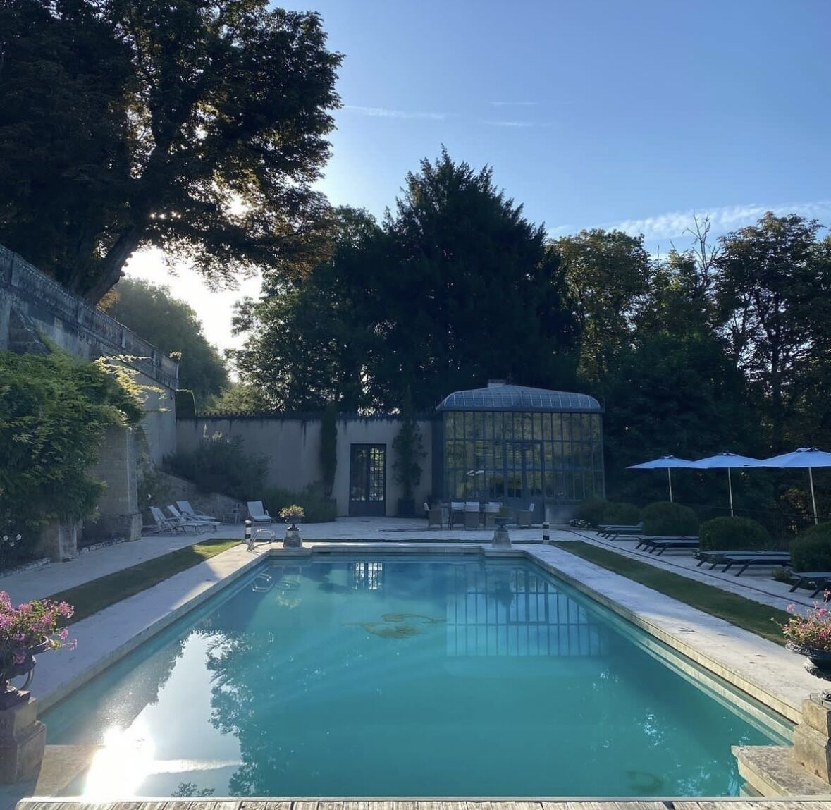 A rectangular pool with clear water, surrounded by a paved deck. There are lounge chairs and umbrellas, and a glass greenhouse structure in the background.