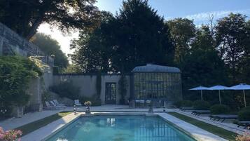 A rectangular pool with clear water, surrounded by a paved deck. There are lounge chairs and umbrellas, and a glass greenhouse structure in the background.