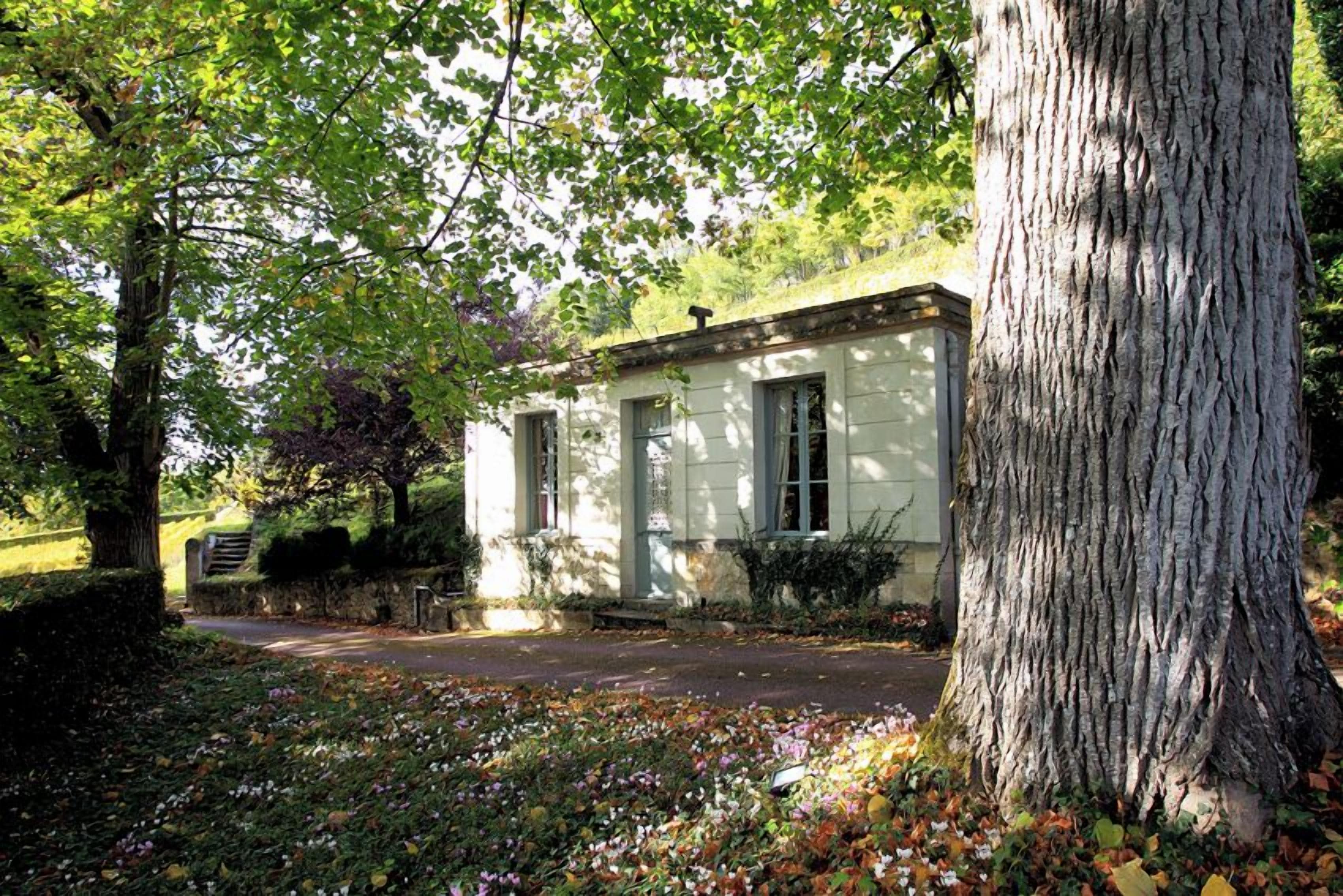 Un bâtiment blanc, doté de fondations en pierre, entouré d’arbres et d’un jardin où les feuilles tombées jonchent le sol.