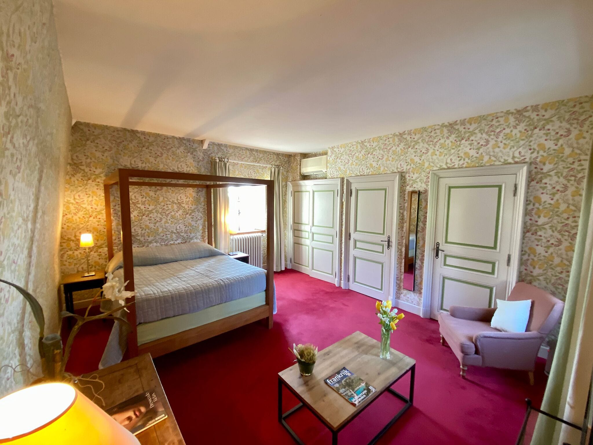 Ein Schlafzimmer mit einem Himmelbett, einem hölzernen Nachttisch, einem Beistelltisch mit Lampe, einem kleinen Tisch mit Blumen und einem Sessel.