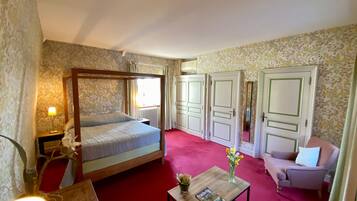 Ein Schlafzimmer mit einem Himmelbett, einem hölzernen Nachttisch, einem Beistelltisch mit Lampe, einem kleinen Tisch mit Blumen und einem Sessel.