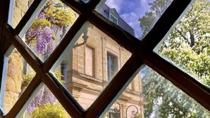 Visão através de uma janela com padrão geométrico, revelando um edifício de arquitetura clássica e um jardim bem cuidado.