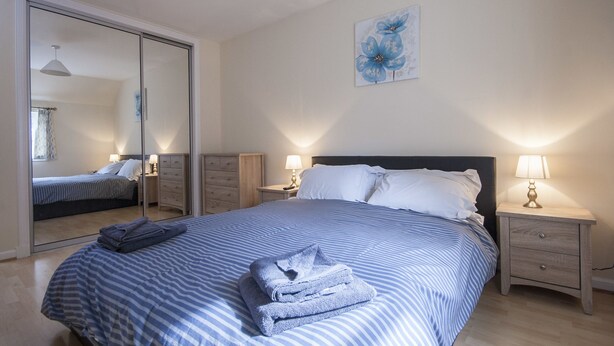 Ein Schlafzimmer mit einem großen Bett, einem Nachttisch mit Lampe, einem Spiegel und einem an der Wand hängenden Kunstwerk.