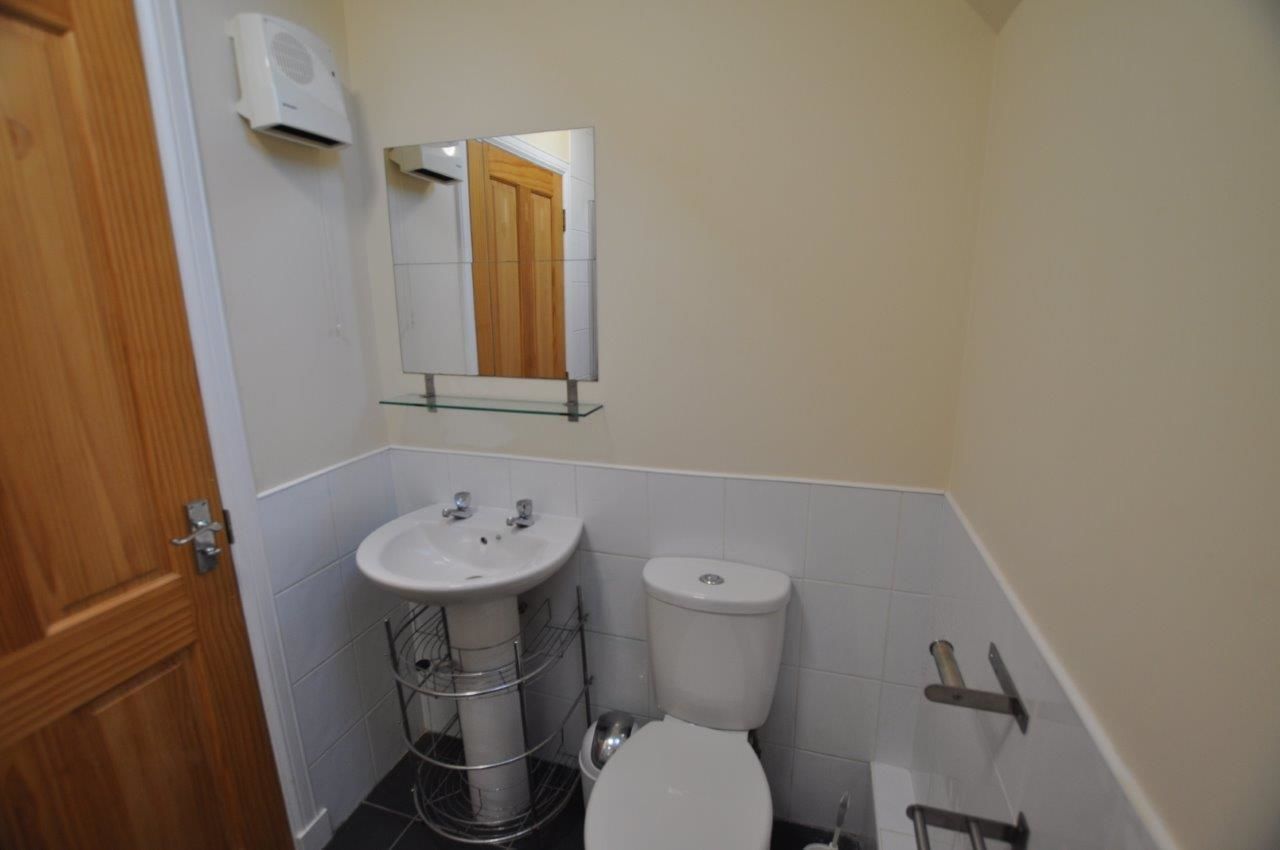 Un baño pequeño con puerta de madera, azulejos blancos, un lavamanos blanco con un espejo encima y un inodoro blanco.