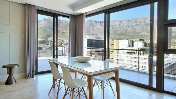 Un comedor moderno con una mesa de vidrio y sillas blancas, ventanas amplias con vistas a la montaña y una pequeña mesita auxiliar.