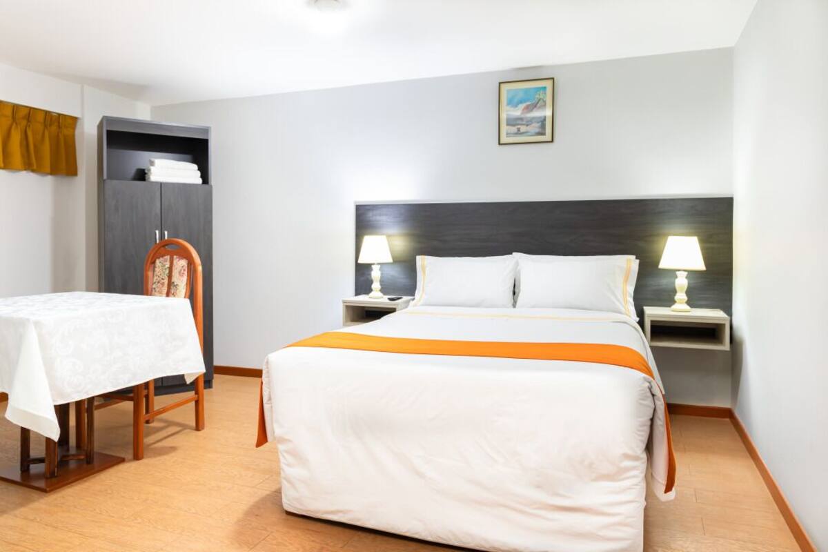 A hotel room with a bed, two bedside tables with lamps, a small table with a tablecloth, a wardrobe, and a painting on the wall.