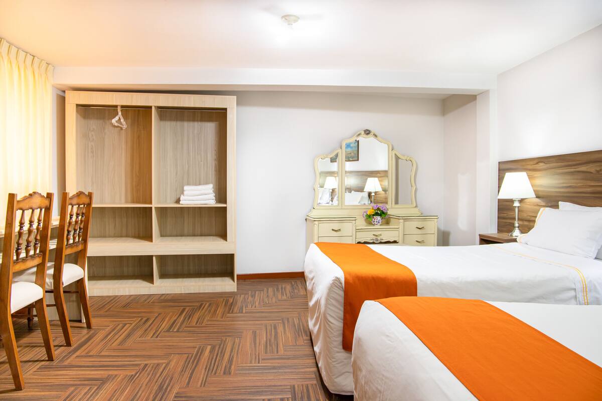 A hotel room with two beds, a wardrobe, a desk with chairs, a mirror, and a nightstand with a lamp.