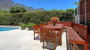 An outdoor patio with a wooden table and benches, surrounded by a pool and a mountain backdrop. - Vetho Villa (Cape Town)