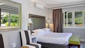 A hotel room with a bed, bedside table, lamp, and a view of greenery through the window. - Vetho Villa (Cape Town)