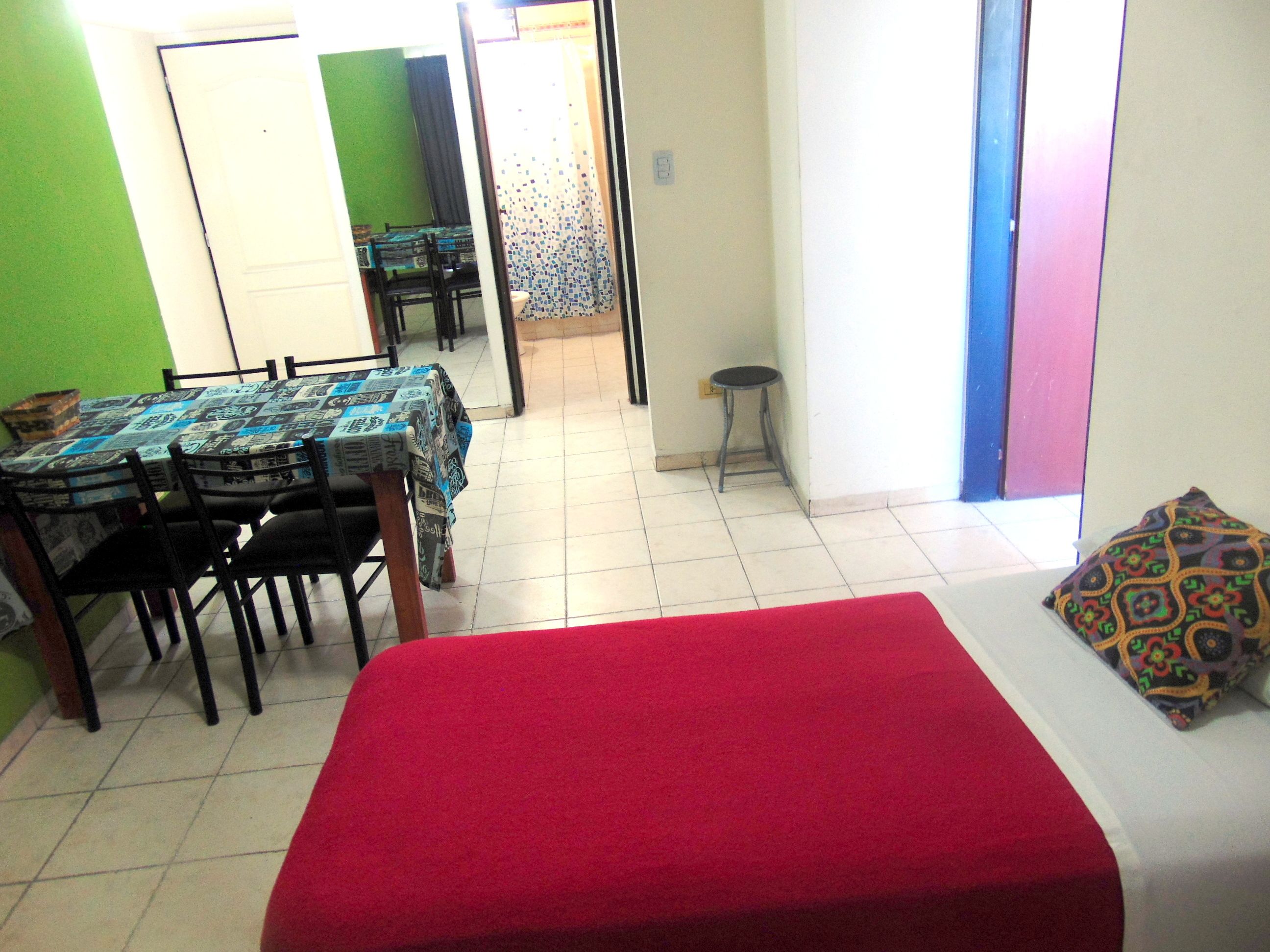 A room with a bed, a dining table with chairs, a small stool, and a colorful pillow.