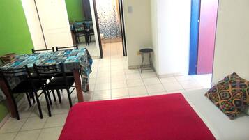 A room with a bed, a dining table with chairs, a small stool, and a colorful pillow.
